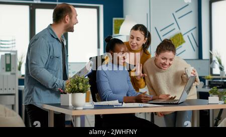 Smiling successful employee with great work results receiving congratulations from coworkers at desk. Woman working on laptop happy with business turnover celebrates with team in startup office. Stock Photo