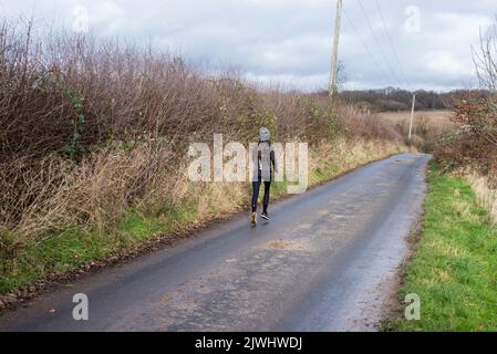 A female runs along a country lane by a tall hedgerow on a tarmac road in the countryside of North London in England Stock Photo