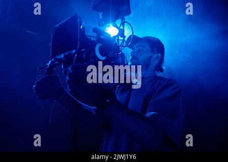 Young man in casualwear holding video camera while shooting commercial in studio lit with blue light and full of smoke Stock Photo