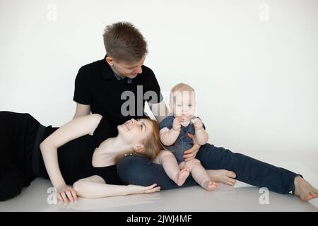 Portrait of young wonderful family in dark clothes with plump cherubic baby infant toddler sitting on white background. Stock Photo