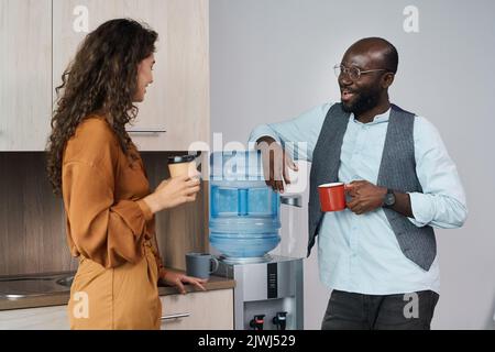 Happy young intercultural office workers chatting and having tea and coffee at break while standing by water dispenser with gallon Stock Photo