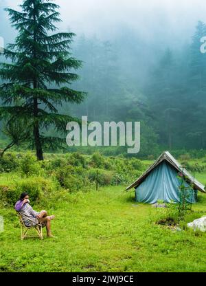 July 7th 2021. A man sipping on a hot tea cup in the meadows with a camp amid deodar tree forest and mountains in the background. Uttarakhand India. Stock Photo