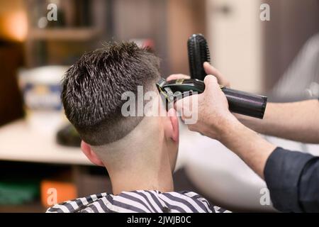 Unrecognizable male barber using electric razor to shave dark hair of man during work in barbershop Stock Photo