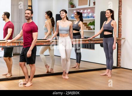 Full length of young positive fit barefooted male and female athletes in sportswear standing near mirror and smiling during barre class in spacious st Stock Photo
