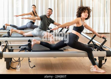 Group of barefoot women and man in activewear spreading arms and looking away while doing stretching exercise on reformers during pilates training in Stock Photo