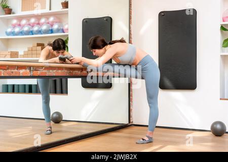 Woman doing leg stretching exercises in a barre in a gymnasium in a health and fitness concept reflected in a wall mirror Stock Photo