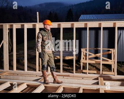 Male developer building wooden frame house. Portrait of man standing on construction site in safety orange helmet, inspecting quality of work. Stock Photo