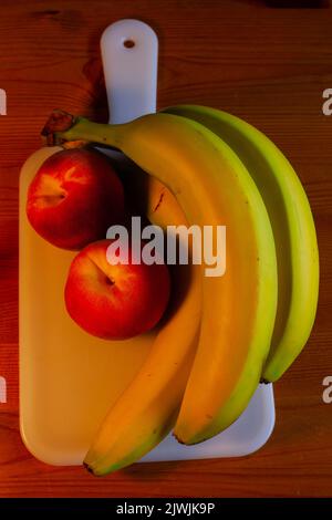 A small bunch of yellow bananas and two peaches are on a white plastic cutting board. Stock Photo