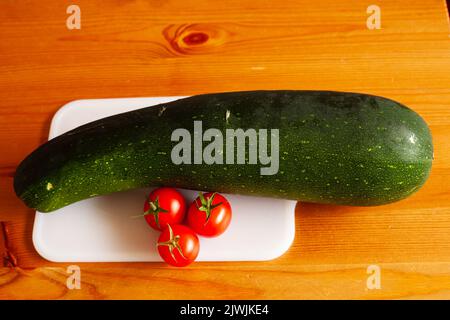 a zucchini lies on a white cutting board and on a brown wooden table with three cherry tomatoes. Stock Photo