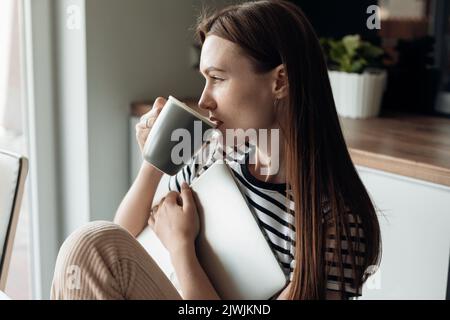 Young dreaming and contemplating female drink tea and holding white laptop in the light kitchen home. Coffee break, rest Stock Photo