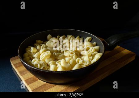 A frying pan with pasta stands on a cutting board made of wood. Food on a black background Stock Photo