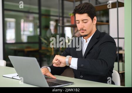 Busy young latin male entrepreneur or employee in formal wear sitting in office and working on laptop, checking the time from a watch on his wrist, time to start online conference or virtual meeting Stock Photo