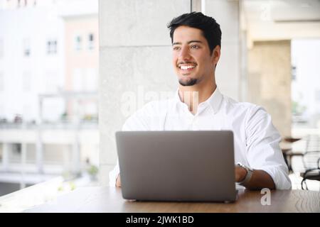 Handsome young dark-haired latin man in white smart casual shirt working on a laptop on the terrace of summer cafe during break. Developer, programmer, copywriter enjoying remote work outdoors Stock Photo