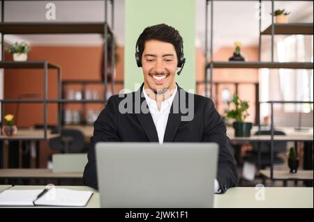 Young confident male remote consultant wearing headset using laptop computer to virtual connection with customers or employees, customer service support manager talking at virtual meeting Stock Photo