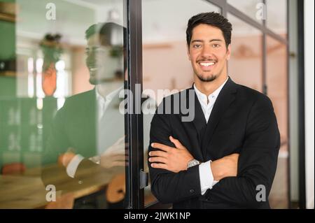 Successful cheerful latin confident young bearded brunet adult male entrepreneur businessman professional in formal wear standing with arms crossed at the modern office, looking at camera Stock Photo
