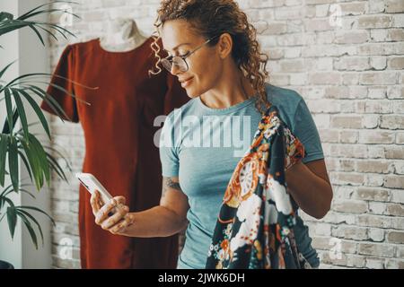 Modern female seamstress calling and using mobile phone at the workshop working with clothes and fashion business. One happy woman people work as a ta Stock Photo