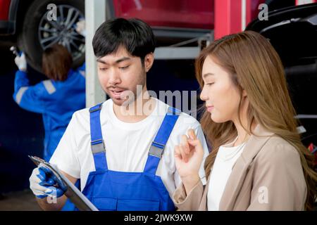 Young mechanic talking to customer asian woman about car with repairing in garage, auto service, technician man explaining problem of vehicle and look Stock Photo