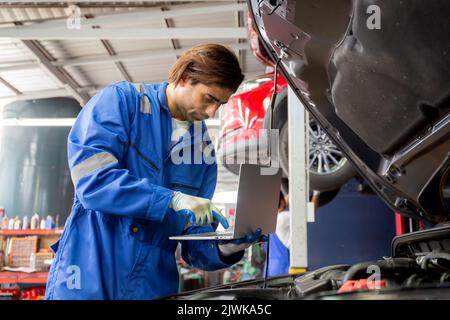 Young man is mechanic using laptop computer checking engine of car in the garage, worker examining and diagnostic automobile, technician and maintenan Stock Photo