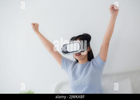 Young asian woman wearing vr headset playing game with excited in the bedroom at home, female using virtual reality or metaverse innovation for simula Stock Photo