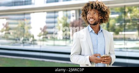Panoramic photo of successful confident elegant mixed race curly haired man, wearing stylish formal clothes, stands outdoors near business center, uses his smartphone, looks away, dreaming, smiling Stock Photo