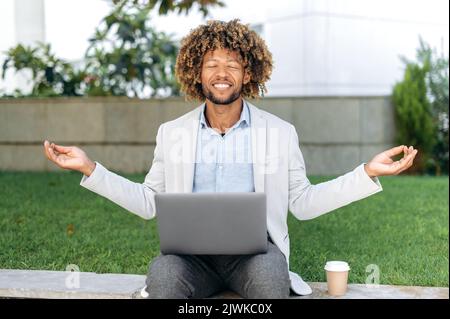 Break during work. Calm relaxed curly haired hispanic or brazilian man, office employee, sits outdoors near the business center, meditates, relaxes, closed his eyes, takes care of mental health, smile Stock Photo