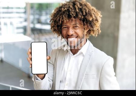 Photo of a joyful friendly mixed race curly haired man, stands outdoors in stylish clothes, shows smart phone with empty white mockup screen, space for advertising, looks at camera, smiles happily Stock Photo