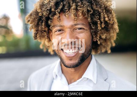 Close-up photo of a handsome attractive confident curly haired latino or brazilian man, stylishly dressed, mixed race successful executive, standing outdoors, looking at camera, smiling friendly Stock Photo