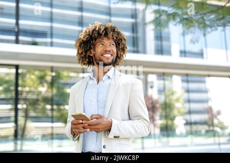 Typing message. Successful confident elegant mixed race curly haired man, wearing stylish formal clothes, stands outdoors near business center, uses his smartphone, looks away, dreaming, smiling Stock Photo