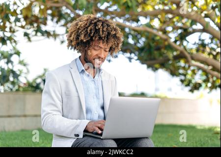 Positive smart successful brazilian or latino curly man, company ceo, programmer, content manager, sitting outdoors with laptop, working online on a project, browsing the internet, smiling happily Stock Photo
