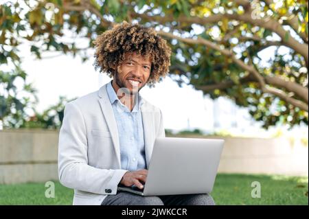 Portrait of positive smart successful mixed race curly man, company ceo, web developer, content manager, sit outdoors with laptop, working online on a project, browsing internet, looks at camera,smile Stock Photo