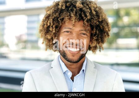 Close-up photo of a confident successful elegant curly haired mixed race man, brazilian or latino entrepreneur, company executive, stand outdoors, looking at camera, smiling friendly Stock Photo