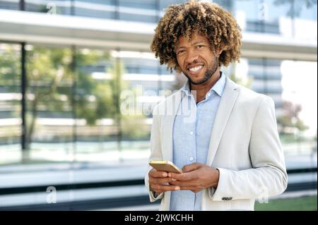 Online messaging. Successful confident elegant latino or brazilian curly haired man, wearing stylish formal clothes, stands outdoors near business center, uses his smartphone, looks at camera, smiles Stock Photo