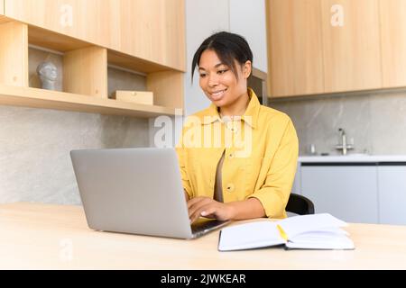 Portrait of a young African American woman sitting at the desk with a laptop, typing an email, messaging friend, chatting, dating online with a pleasant smile, long-distance relationships concept Stock Photo