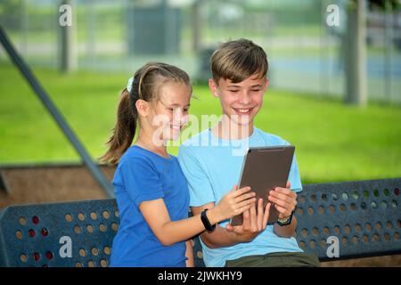 Happy teenager children, girl and boy looking at screen of digital tablet, reading, studying or gaming sitting on bench outdoors on summer sunny day. Stock Photo