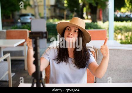 Attractive woman outside sitting with tripod and smartphone,making video call or providing online lesson.She showing thumbs up. Stock Photo