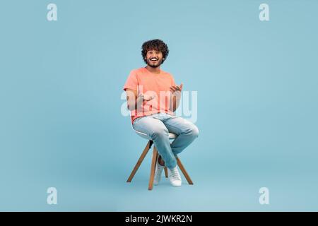 Cheerful eastern guy sitting on chair over blue background Stock Photo