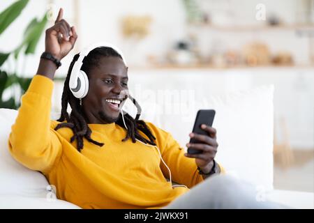 Carefree Black Guy Relaxing On Couch With Smartphone And Wireless Headphones Stock Photo