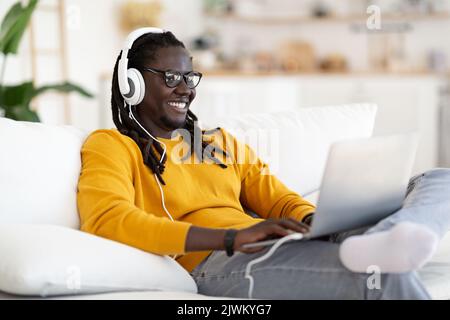 Smiling Young Black Man Wearing Wireless Headphones Relaxing With Laptop At Home Stock Photo