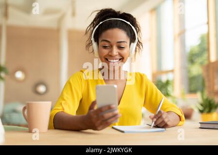 Remote learning. Cheerful black lady in wireless headphones sitting at desk using smartphone and writing in her notebook Stock Photo