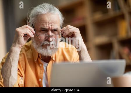 Concentrated old man using notebook at home, touching his eyeglasses Stock Photo