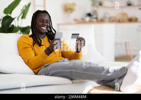 Online Shopping. Happy Young Black Man Using Smartphone And Credit Card Stock Photo