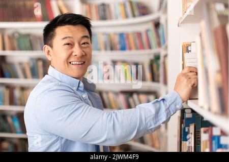 Cheerful Asian Man Taking Book From Bookshelf In Modern Library Stock Photo