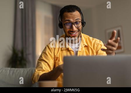 Millennial black man in headset having web call, using laptop computer, sitting on couch, talking at webcam at home Stock Photo