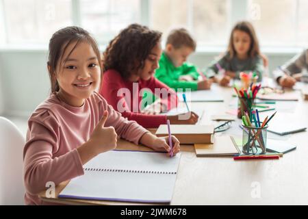 Asian Schoolgirl Gesturing Thumbs Up Smiling Taking Notes In Classroom Stock Photo