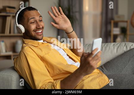 Happy young black man in headset communicating on smartphone, waving at webcam, sitting on sofa at home Stock Photo