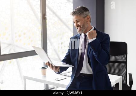 Portrait Of Overjoyed Middle Aged Businessman Celebrating Success With Laptop In Office Stock Photo