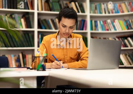 Serious woman sitting at desk in library using laptop and taking notes in notebook, studying online, copy space Stock Photo