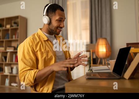 Happy young black man using laptop, wearing headphones, having video chat at home, copy space Stock Photo