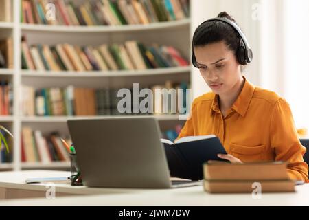 Focused female student reading book and using laptop computer, studying online, sitting in library, copy space Stock Photo