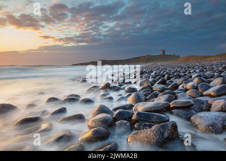 Breaking waves on rocks below Dunstanburgh Castle at sunrise, Embleton Bay, near Craster, Northumberland, England, United Kingdom, Europe Stock Photo